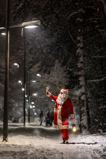 Santa Claus walking outdoors and enjoying wonderful snowy December night in city park before Christmas and holidays. He is carrying lantern and bag full of gifts.
