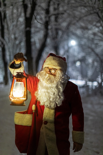 Santa Claus walking outdoors and enjoying wonderful snowy December night in city park before Christmas and holidays. He is carrying lantern and bag full of gifts.