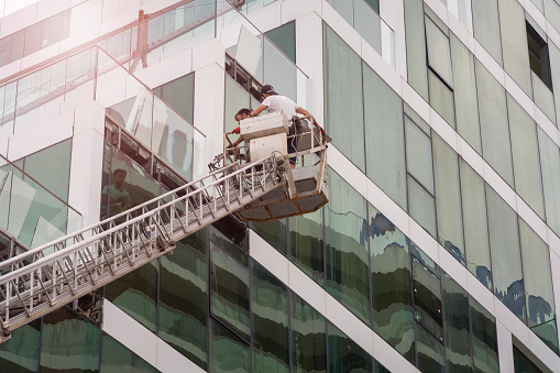 Batumi, Georgia - August 1 2022: Male workers on the stairs, the boom of the lifting machine are repairing something on the facade of a modern glass building