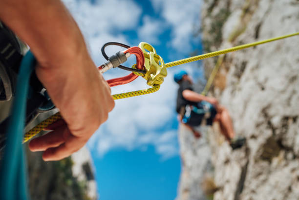 primer plano del dispositivo belay con un niño en el muro de escalada del acantilado. él colgado de una cuerda en un arnés de escalada y su compañero asediándolo en el suelo. imagen de personas activas y concepto deportivo - rock climbing fotos fotografías e imágenes de stock