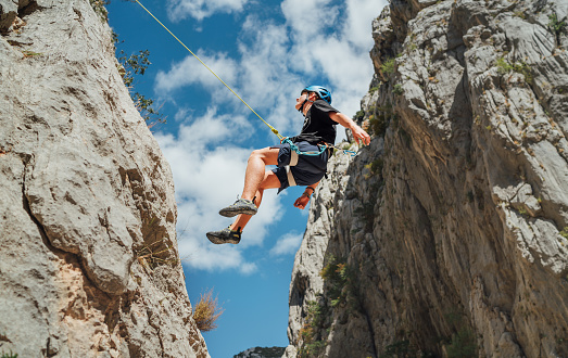 Climber teenager boy in protective helmet abseiling from vertical cliff rock wall using rope Belay device, climbing harness in Paklenica canyon in Croatia. Active extreme sports time spending concept