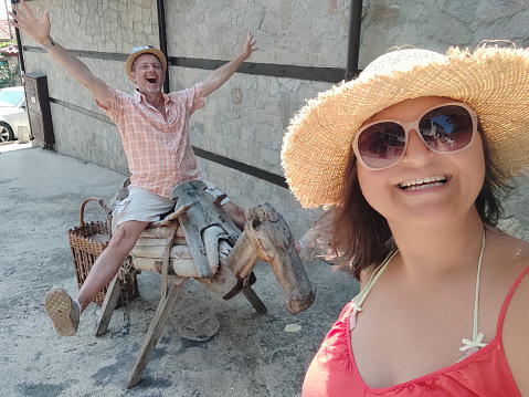 A cheerful woman in a red dress, sunglasses and a knitted straw hat takes a selfie on a quiet street in a small resort town. Behind her, a cheerful man in an orange plaid shirt, sunglasses, and a knitted straw hat rides a wooden donkey.