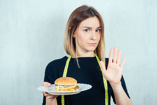young and attractive blond woman in black T-shirt and measuring tape is holding a high-calorie burger on the plate and shows a hand gesture stop. concept of refusing harmful fast food and diet.