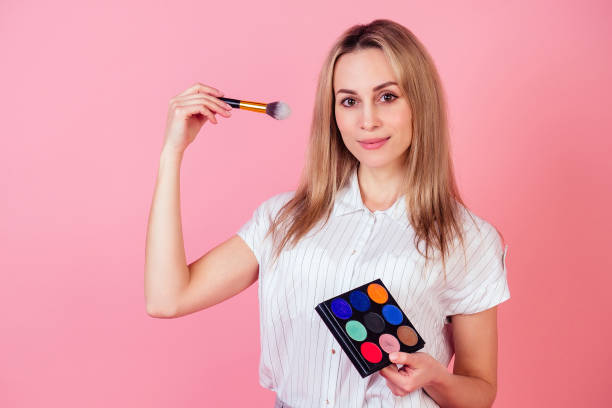 attraktive blonde visagiste make-up artist frau mit einem make-up-pinsel und einer palette von hellen lidschatten auf einem rosa hintergrund im studio. konzept der kosmetik und hautpflege - luggage hold stock-fotos und bilder