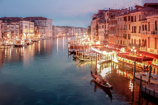 View of Grand Canal in downtown Venice just after sunset, with magic light illuminating the scene. Streetlights shining on piers and canal banks, while gondolas move slowly on water.
