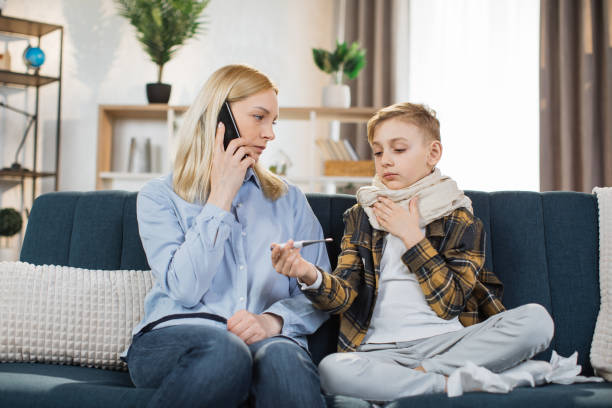 Boy with scarf around neck sitting on sofa at home with a thermometer after measuring the temperatur Sad sick teen boy with scarf around neck sitting on sofa at home with a thermometer after measuring the temperature, while his worried young mother talking with doctor by phone. Flu, cold concept. temperatur stock pictures, royalty-free photos & images