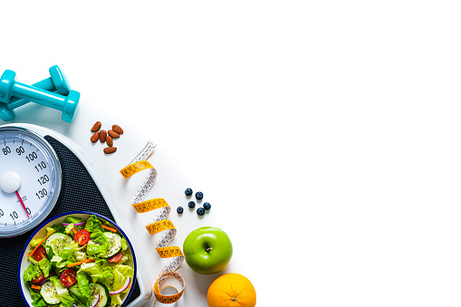 Overhead view of a composition of healthy fruits, salad plate, weight scale, tape measure and dumbbells shot on white background. The composition is at the left of an horizontal frame making a border and leaving copy space.  High resolution 42Mp studio digital capture taken with SONY A7rII and Zeiss Batis 40mm F2.0 CF lens