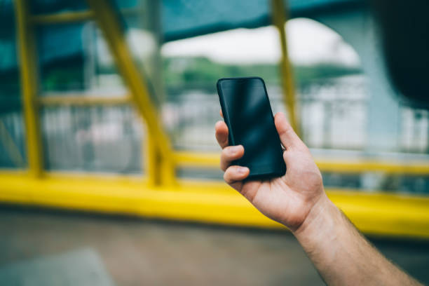 Crop hand of faceless person holding black cellphone with empty screen in glass covered pedestrian bridge on city street  on blurred background