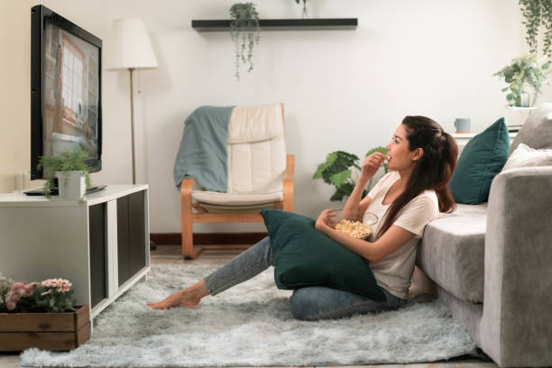 自宅のリビングルームでポップコーンを食べながら、テレビでオンデマンドで映画ビデオを見ている床に座って幸せな女性。 - streaming media service ストックフォトと画像