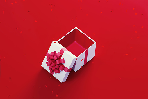 White gift box tied with red ribbon sitting under red falling confetti on red background. Horizontal composition with copy space. High angle view. Great use for Christmas and Valentine's Day related gift concepts.