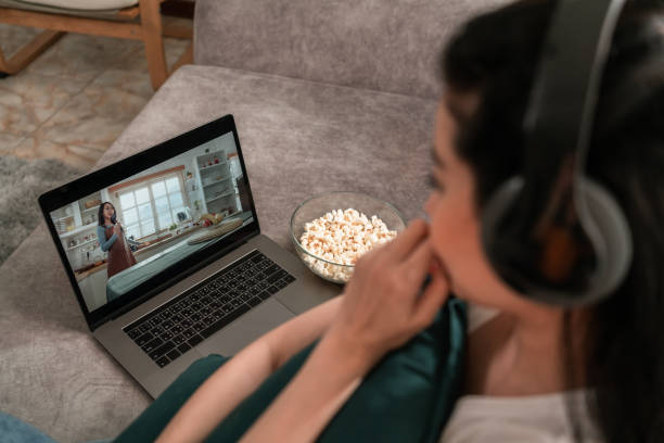 肩越しに、自宅のリビングルームでラップトップでオンデマンドで映画ビデオを見ているヘッドフォンを着用した女性が見る - streaming media service ストックフォトと画像