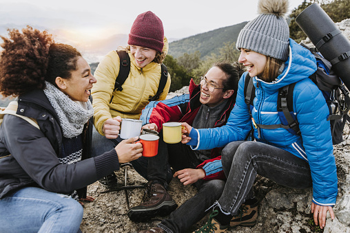 Group of four hikers with backpacks drinking hot coffee on the top of the mountain - Happy friends hiking in nature together - Trekking lifestyle and adventures concept