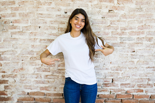 Beautiful hispanic woman pointing to her design logo at her mockup white t-shirt standing in front of a brick wall outdoors
