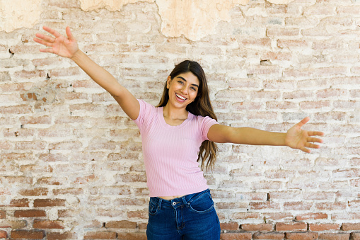 Excited cheerful young woman opening her arms and celebrating or ready to give a hug while laughing in front of a brick wall