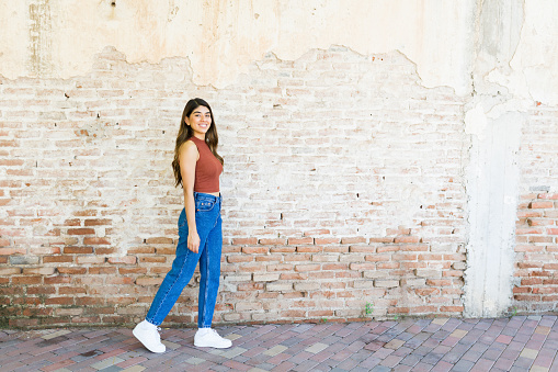 Full length of a beautiful smiling woman on casual clothes walking outside beside a brick wall with copy space