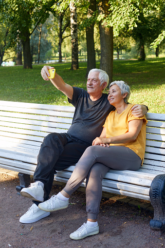 Happy senior spouses take selfie picture sit on bench in summer park. Grey-haired couple look at cell phone screen make videocall outdoor use modern tech, enjoy online fun together spend time outside