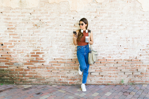 Full length of a latin young woman leaning on a brick wall while drinking coffee and texting on the phone while waiting to go to work