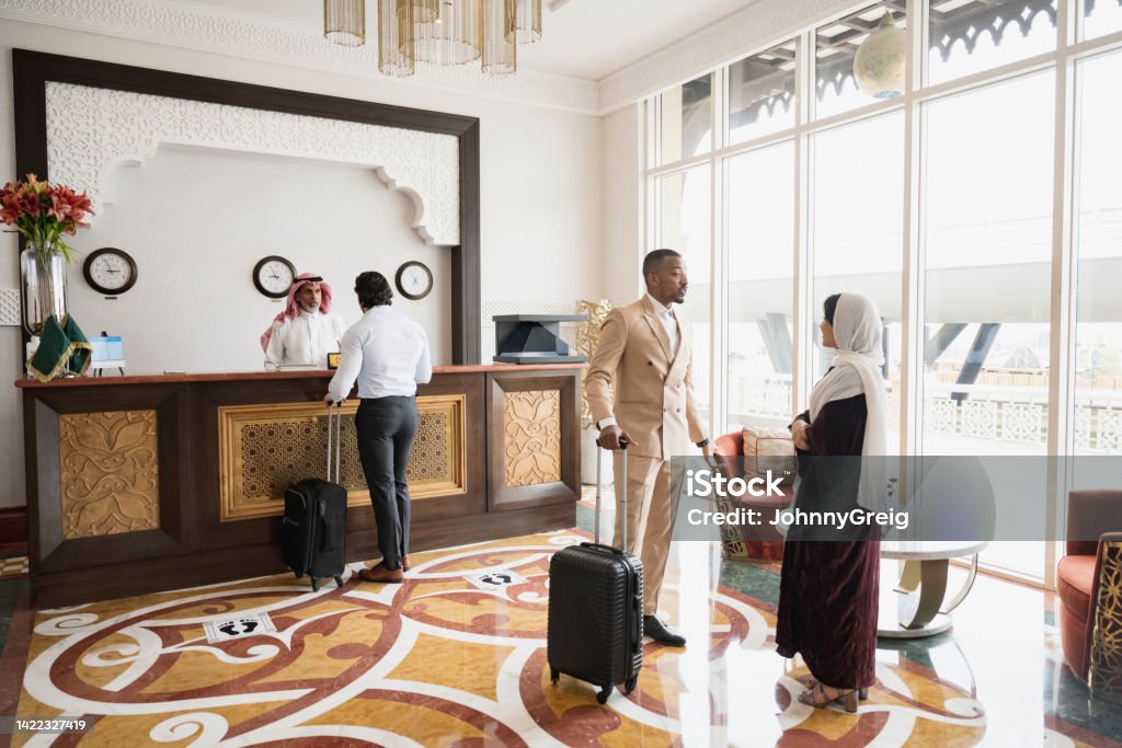 Hotel guests standing in lobby and at reception desk Full length view of business traveller talking with receptionist and vacationing couple with luggage waiting to check-in at small boutique hotel in Riyadh. Hotel Reception Stock Photo