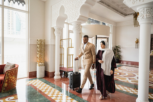 Full length view of early 30s man in suit with wheeled luggage and mid 20s woman in abaya and hijab walking through elegant lobby with Islamic design features.