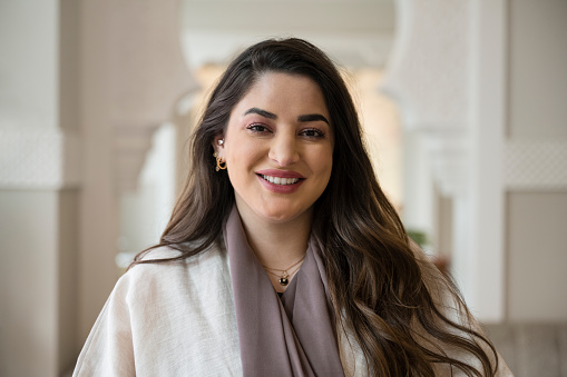 Head and shoulders view of Saudi woman with long brown hair standing in lobby of Riyadh hotel with traditional Islamic design and smiling at camera.