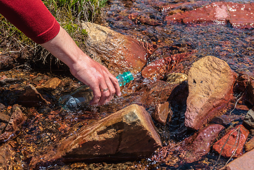Man replenishes water from mountain stream in bright sun. Tourist fills bottle with mineral water in sunny day. Hand with bottle in clear water stream in sunlight. Replenishment of water during hike.
