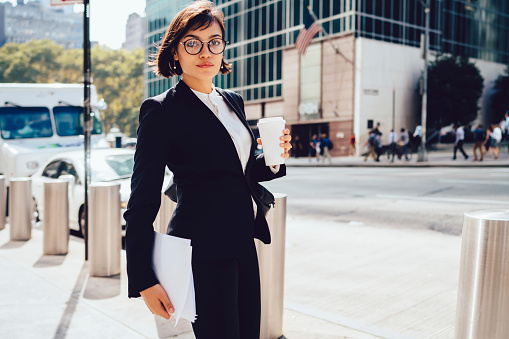 Serious elegant businesswoman in formal wear and eyeglasses standing on street of New Yor City with takeaway coffee and documents