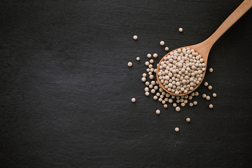 White pepper or peppercorns in wooden spoon on black dark background , top view , flat lay.