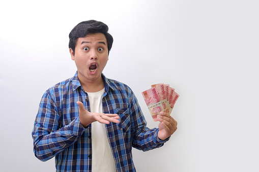 Portrait of excited Asian man in blue plaid shirt standing against white background, showing one hundred thousand rupiah while making surprise hand gesture. Financial and savings concept.