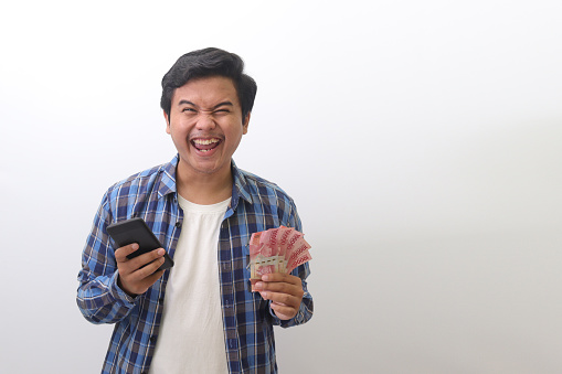 Portrait of excited Asian man in blue plaid shirt standing against white background, showing one hundred thousand rupiah while using mobile phone. Financial and savings concept.