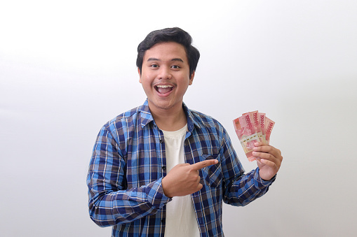 Portrait of excited Asian man in blue plaid shirt standing against white background, showing one hundred thousand rupiah while pointing to the side. Financial and savings concept.