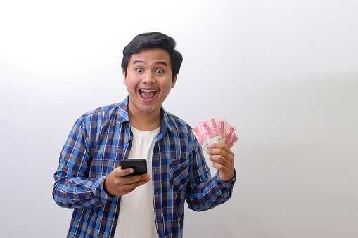 Portrait of excited Asian man in blue plaid shirt standing against white background, showing one hundred thousand rupiah while using mobile phone. Financial and savings concept.