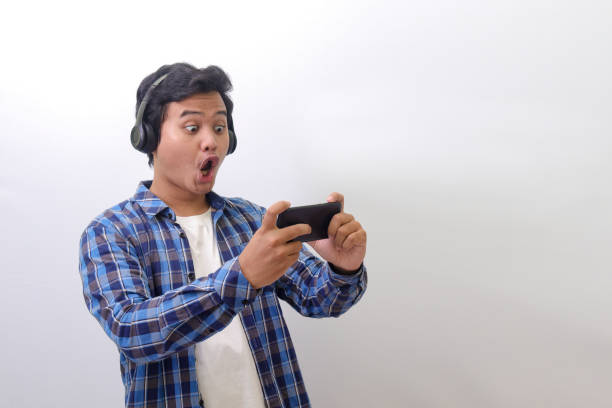excited Asian man using wireless headphone, playing games on his mobile phone by tilting the screen stock photo