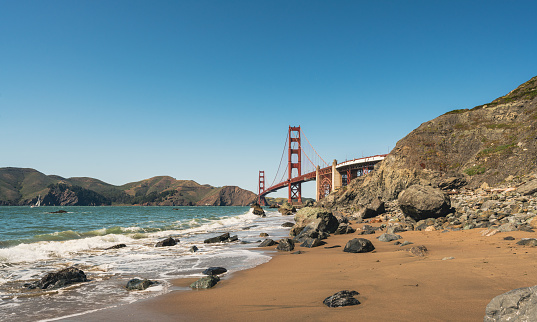 The Golden Gate Bridge in San Francisco seen a sunny morning at the beach in California, USA. Waves from the Pacific Ocean splashing towards the rocks.