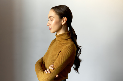Profile view of attractive young office worker standing against white wall with crossed arms and closed eyes, resting, relaxing and meditating using stress relief technique in the middle of work day