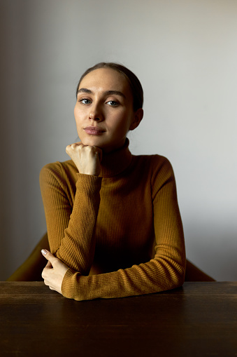 Vertical indoor portrait of serious thoughtful and pensive female therapist looking at camera sitting at table with crossed arms, putting chin on hand, dressed in turtleneck, isolated on beige wall