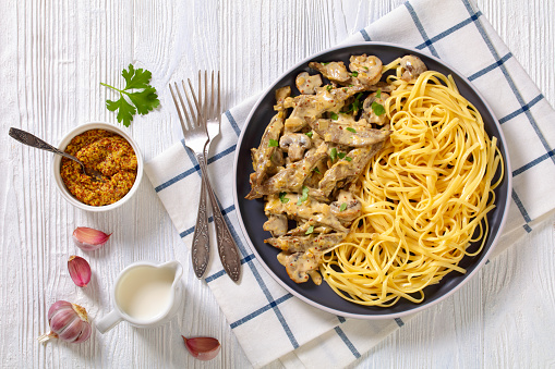 calves liver stroganoff cooked with cream, wholegrain mustard, onion and mushrooms served with spaghetti on plate on white wood table, horizontal view from above, flat lay