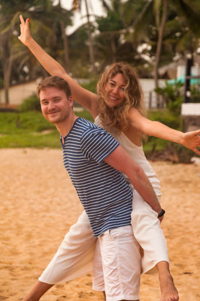 istock Happy lovely couple having fun on tropical sandy beach at palm trees background. Man giving piggyback ride to his woman and laughing. Joyful couple enjoying summer at ocean coastline. Copy text space 1422425166