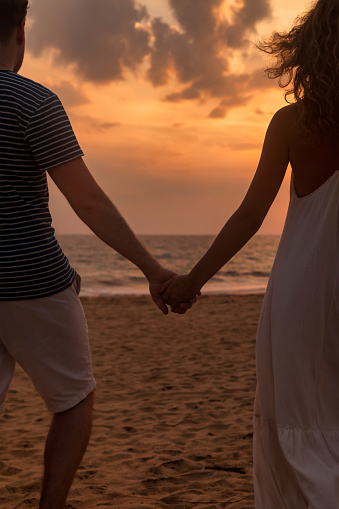 Close up rear view young lovely family couple holding hands, man and woman in casual clothes, walking together sandy beach at ocean sunset background outdoor. Travel vacation concept. Copy text space