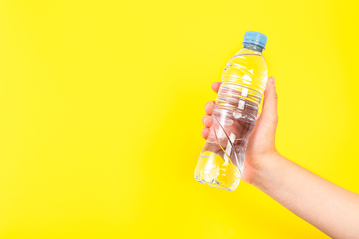 Front view of a young man hand holding a bluish translucent PET  water bottle against a yellow background. The hand is at the lower right corner leaving a useful copy space at the left side of the image.