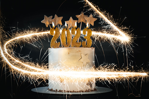 Big golden New Year cake decorated with golden stars and number 2023 on top. Happy New Year 2023. Winter festive background with light trails of sparklers