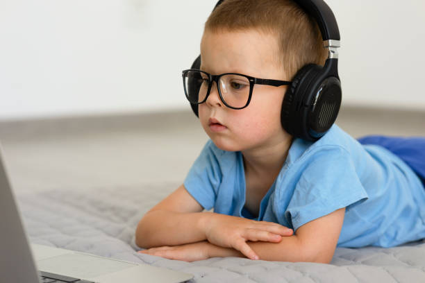 the child lies on his stomach in black headphones and looks into the laptop. the child lies on his stomach in black headphones, glasses and looks into the laptop. Portrait of a little boy with a laptop. The child is looking intently at the screen of the laptop. High quality photo concentrated solar power photos stock pictures, royalty-free photos & images