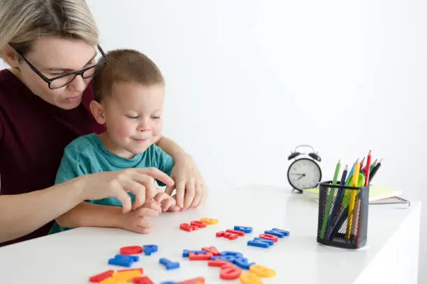 Mom or a teacher shows the child letters and compose words. Mom and son do homework at a white table. Pencils, student, letters, alarm clock, notebooks. High quality photo