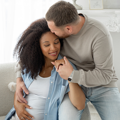 Caucasian man embrace and kiss his pregnant black wife. Pregnant woman and loving husband hugging tummy at home, smile at camera. Square.