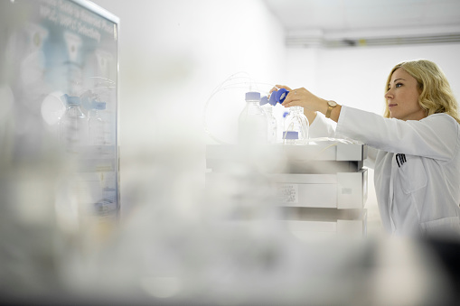 Blonde female employee in a white medical coat seen in a pharmaceutical factory while working on some laboratory research with special equipment during her working hour in a factory for drugs.