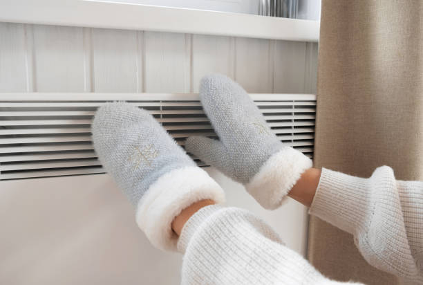 Hands in winter gloves warming up over electric heater. Frozen woman wearing a mittens shaking and freezing for winter cold. Discomfort spending time at home. Girl warming hands on modern radiator. Central Heating Crises. Rise in price of Energy. Global Energy Shortage. International gas supply crisis las manos en guantes de invierno se calientan sobre el calentador eléctrico. mujer congelada con un mitón temblando y congelándose para el frío invernal. incomodidad al pasar tiempo en casa. chica calentando las manos en el radiador moderno.