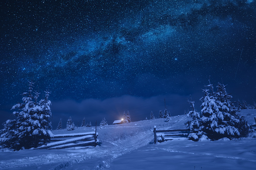 Bright milky way in a night starry sky. Lonely house on a snowy hill with Christmas light.