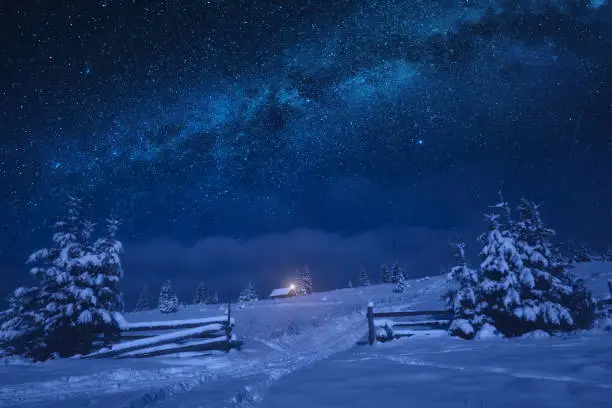 Bright milky way in a night starry sky. Lonely house on a snowy hill with Christmas light.