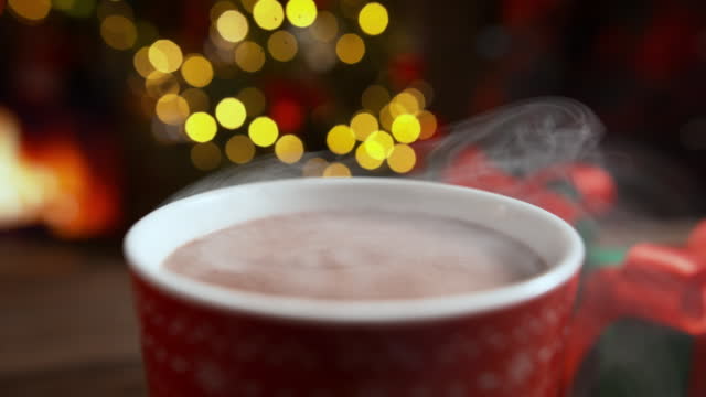 Hot Chocolate with Creamy Foam Steaming in Red Mug with Fireplace and Christmas Lights in the Background