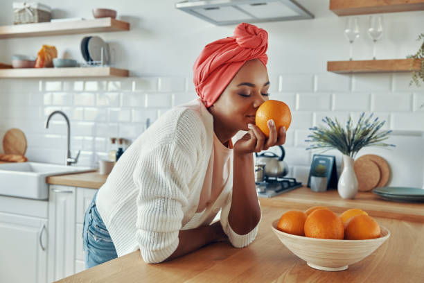 Beautiful African woman in traditional headwear smelling oranges while standing at the kitchen