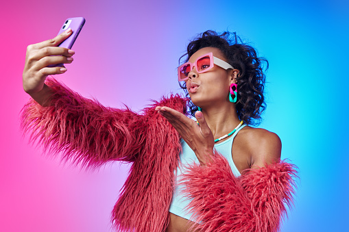 Fashionable African woman in fluffy jacket making selfie and blowing a kiss against colorful background